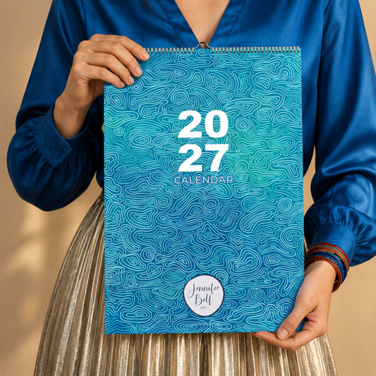 A person in a blue blouse and pleated skirt holds a 2027 spiral-bound calendar with a blue artwork cover. The calendar features the text '2027 CALENDAR' and a circular 'Jennier Bell' logo near the bottom.