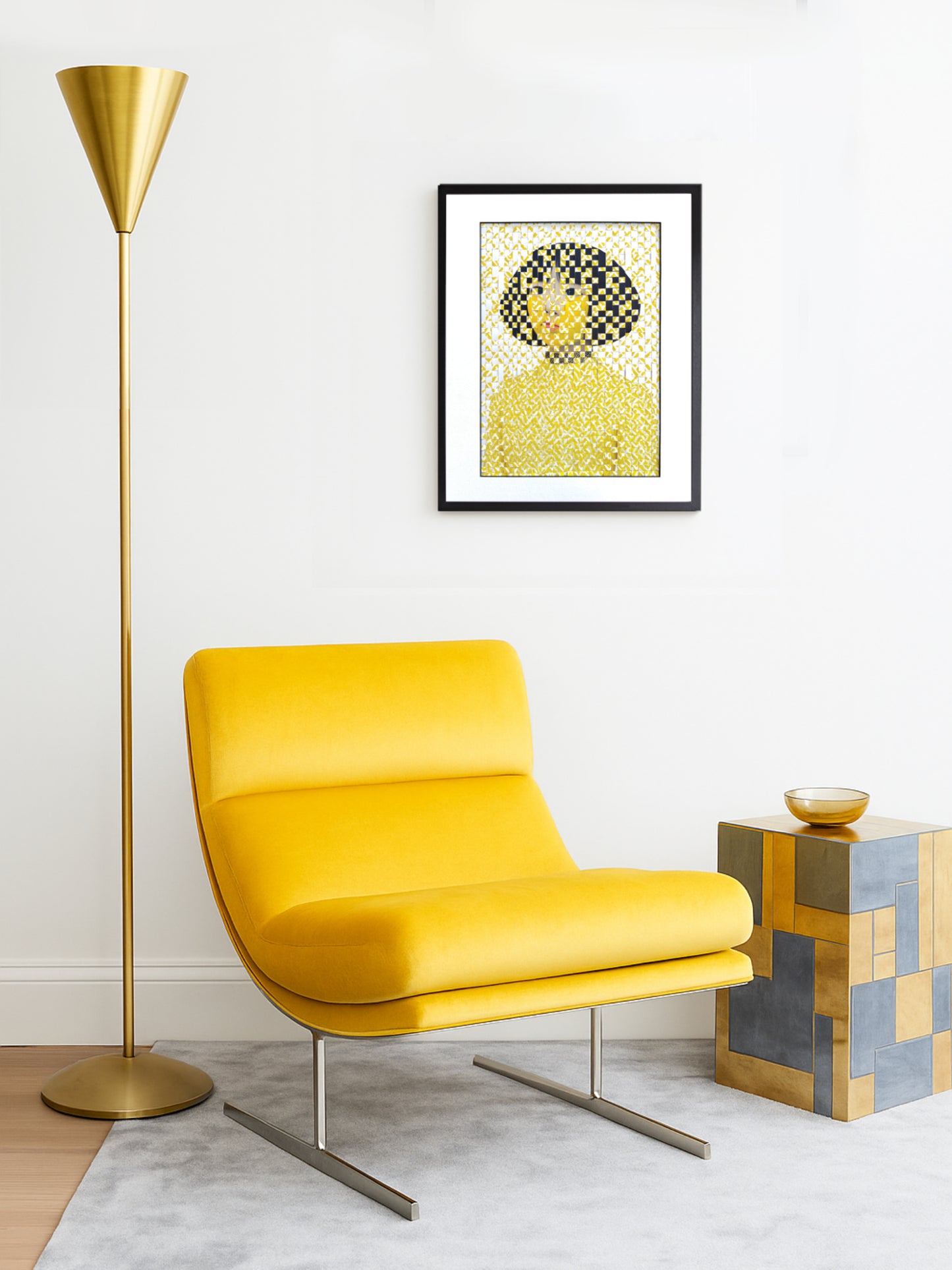 Modern minimalist living room with a mustard yellow lounge chair on a pale grey rug, a tall gold floor lamp to the left, and a small geometric mirrored side table with a gold bowl on top. On the wall above the chair hangs a framed woven portrait artwork in yellow and black tones, which serves as the focal point of the space