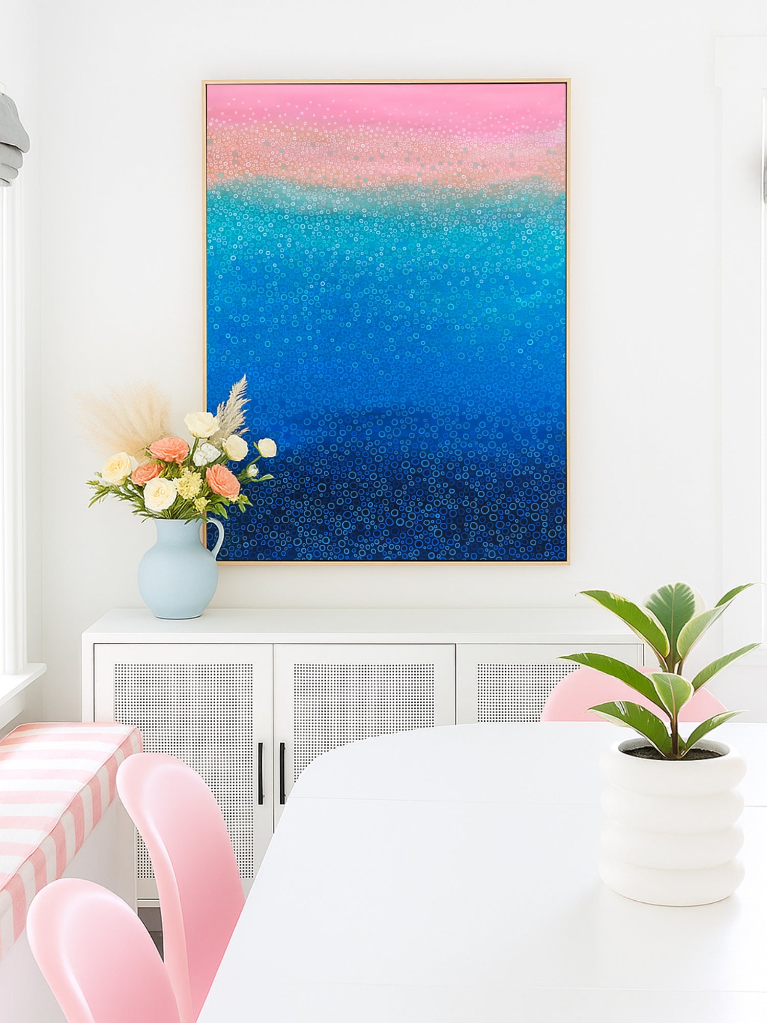 Colourful abstract art with a gradient of pink, blue, and purple above a white cabinet with pink chairs and a plant.