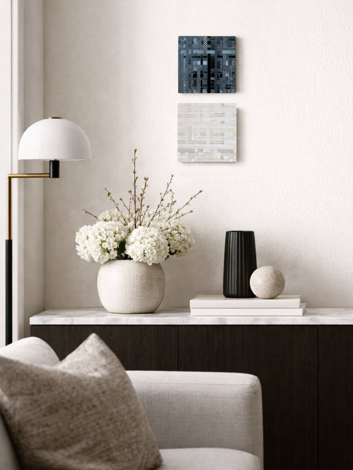 Two small 6×6 inch abstract woven artworks hang vertically on a soft neutral wall in a high end interior, with the dark black and charcoal piece displayed above a lighter ivory and beige piece. A sleek console table with sculptural vases and flowers sits below, creating a refined, minimalist setting that highlights the scale and texture of the artworks.
